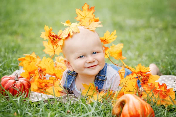 Portrait of cute funny adorable blond Caucasian baby boy with blue eyes in tshirt and jeans romper lying on grass field meadow, yellow autumn fall leaves pumpkins. Halloween, Thanksgiving.
