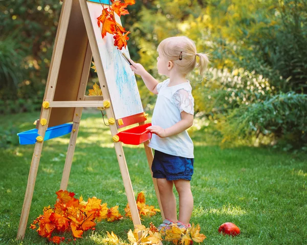 White Caucasian toddler child kid girl standing outside in summer autumn park drawing on easel with markers looking away, playing studying learning, back to school