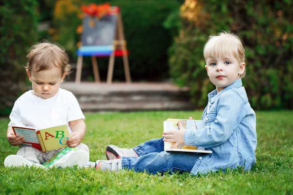 Group of two white Caucasian toddler children kids boy and girl sitting outside in grass in summer autumn park by drawing easel with books, playing studying learning, back to school