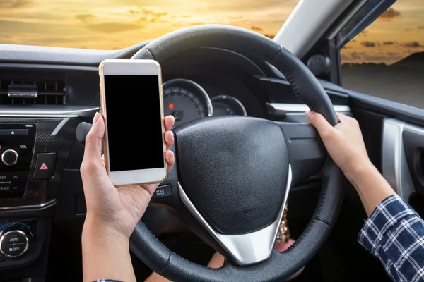 Young woman driver using touch screen smartphone and hand holding steering wheel in a car with sunset background