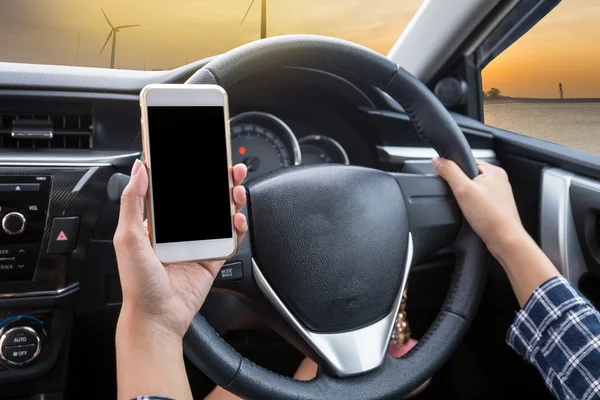 Young woman driver using touch screen smartphone and hand holding steering wheel in a car with wind turbine at sunset background