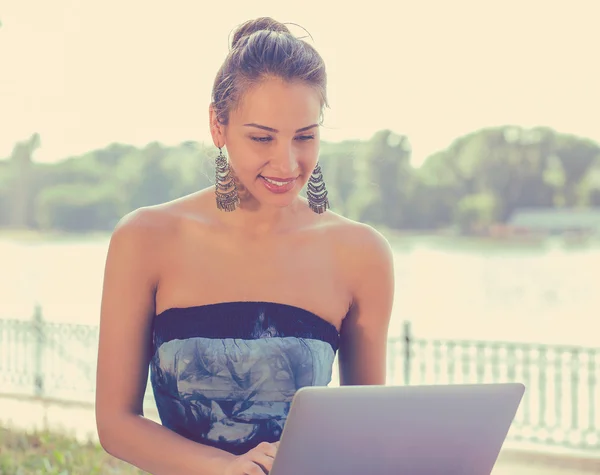 Smiling woman sending emails sitting on grass with computer balanced on knees