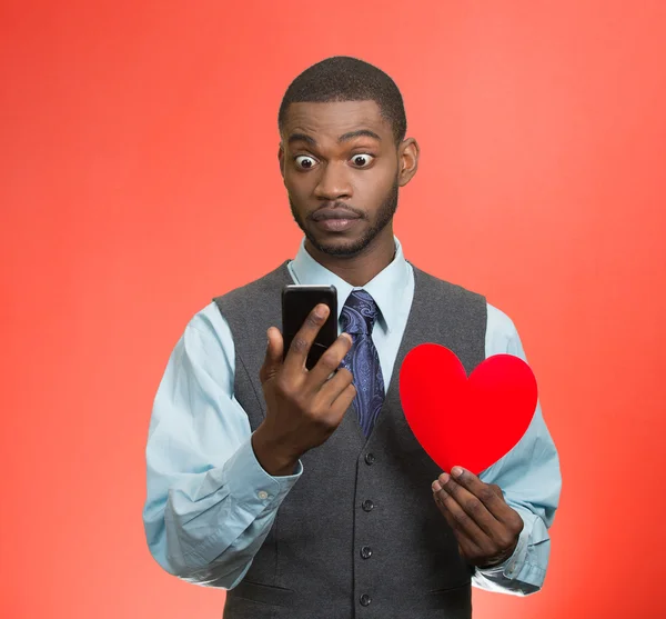 Shocked young man reading breaking news on phone holding red heart