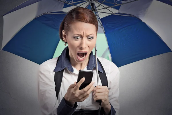 Shocked woman reading breaking news on smartphone holding umbrella