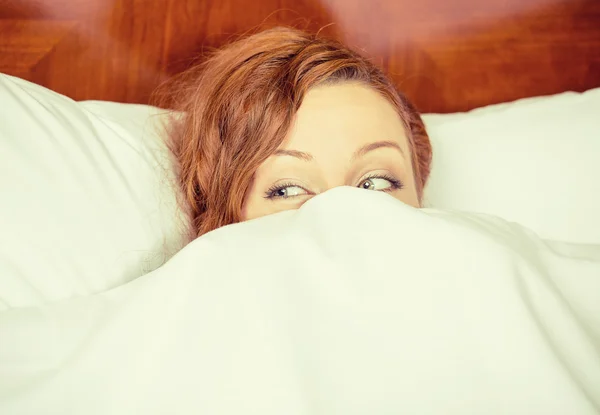 Young shy woman lying in bed hiding under duvet blanket