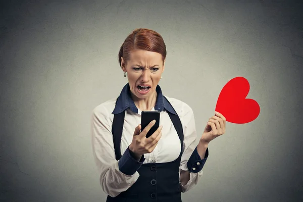 Displeased young woman reading news on smart phone throwing away red heart