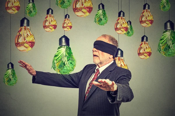 Blindfolded senior man walking through light bulbs shaped as junk food and green vegetables