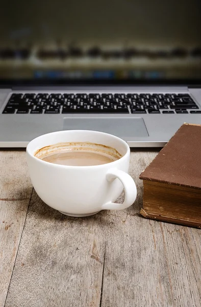 White cup of coffee,old book and laptop on wooden table.