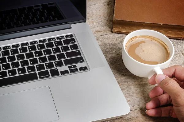 Laptop and cup of coffee in hand on wooden table background.