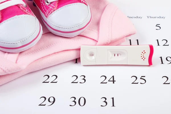 Pregnancy test with positive result and clothing for newborn on calendar, expecting for baby
