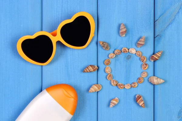 Seashells in shape of sun, sunglasses and sun lotion on blue boards, accessories for summer