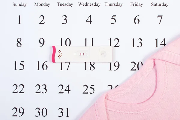 Pregnancy test with positive result and bodysuit for newborn on calendar, expecting for baby