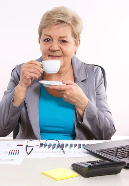 Elderly business woman drinking tea or coffee at desk in office, break at work