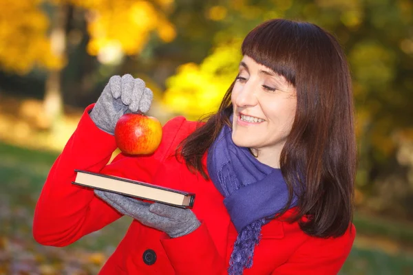 Happy smiling woman holding book and apple in autumn park