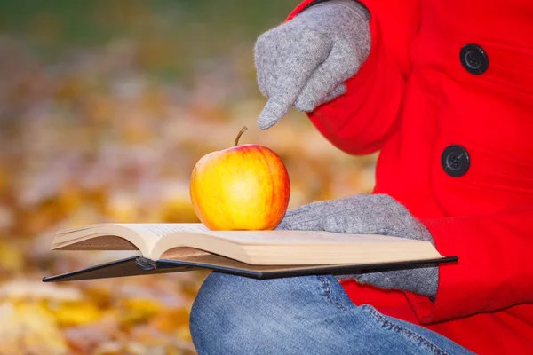 Finger of woman showing book and apple in autumn park