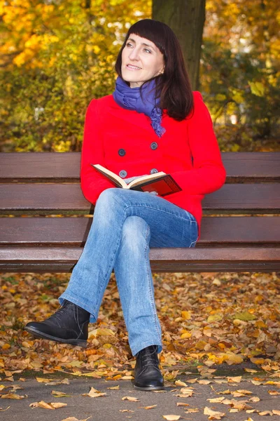 Happy smiling woman sitting on bench reading book in autumn park