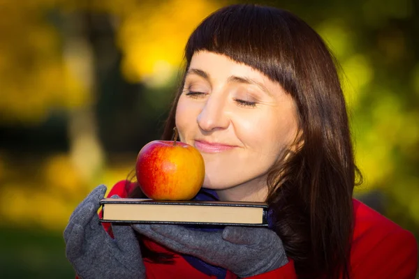 Pensive and dreamy woman holding fresh apple and book in autumnal park, autumn concept