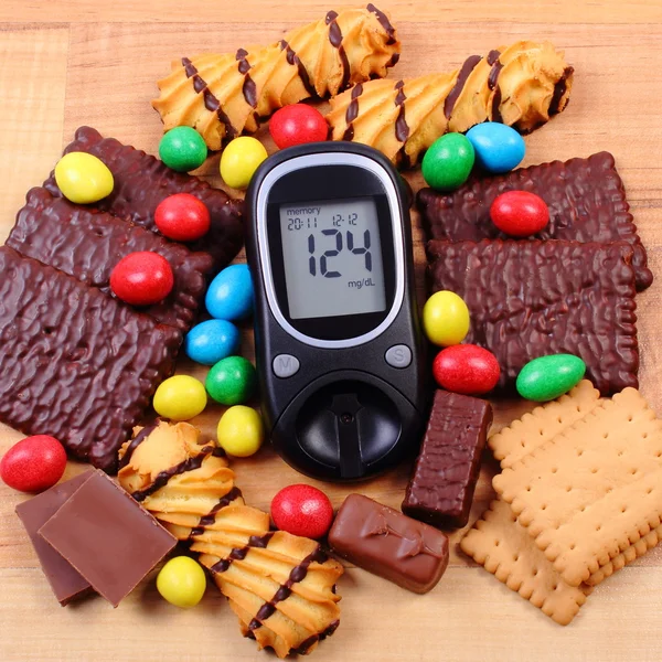 Glucometer with heap of sweets on wooden surface, diabetes and unhealthy food