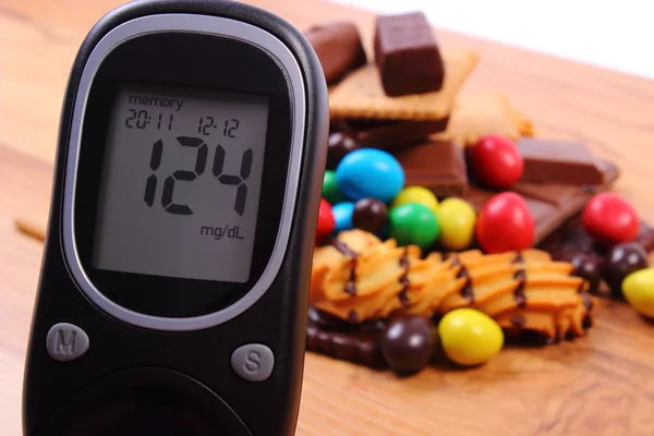 Glucose meter with heap of sweets on wooden surface, diabetes and unhealthy food