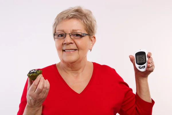 Happy senior woman holding glucometer and fresh cupcake, measuring and checking sugar level, concept of diabetes