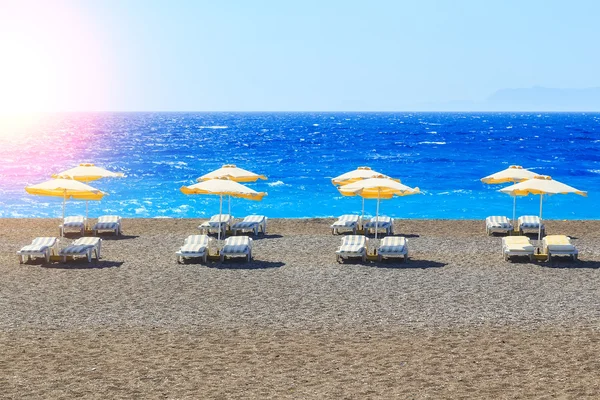 Greece, reed umbrellas and yellow sunbeds on pebble beach at Aegean Sea of Rhodes,