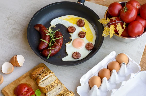 Fried sausages and eggs in a pan served with bread , tomatoes and egg shell on a grey stone board