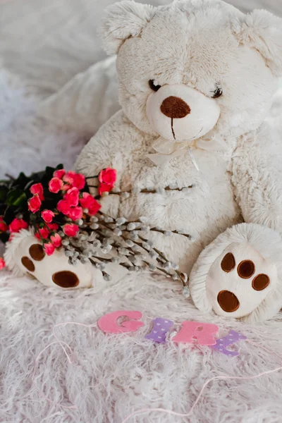 A beautiful soft toy bear sits on the bed with flowers