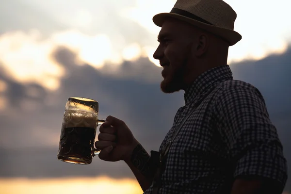 Happy smiling man tasting fresh brewed beer against the sky at sunset. The theme is Oktoberfest, a guy in Bavarian style