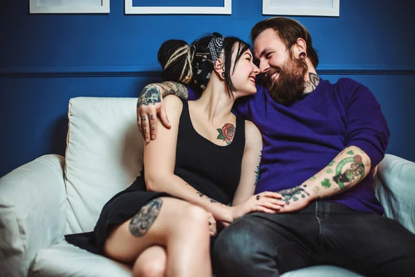 Young couple in love with tattoos hugging and holding hands sitting on sofa