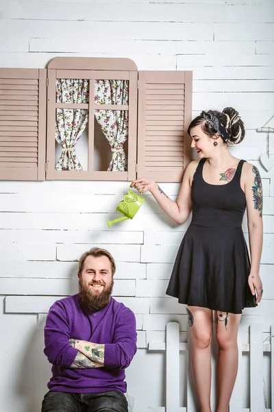 A guy and a girl with tattoos having fun, a man sits on the bench and the girl standing with a watering can