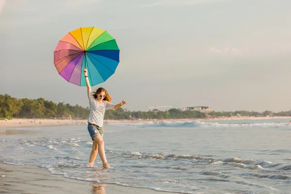 Cheerful young girl with rainbow umbrella having fun on the beach before sunset. Travel, holidays, vacation, healthy lifestyle concept