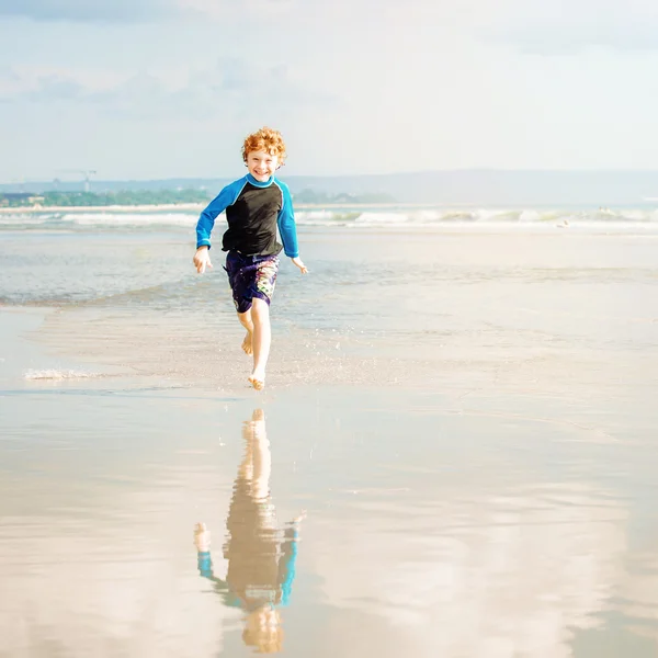 Young boy in swimming shorts and rash vest runs along Bali beach near sunset