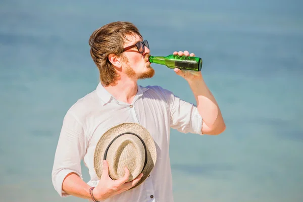 Man wearing hat and sunglasses enjoing beer in a bottle on the beach
