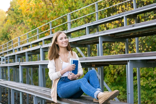 Student sitting on sport tribune with book and smiling at camera