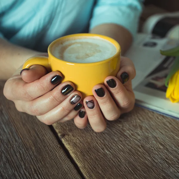 Yellow cup in the hands of women. Gel nail manicures. Phone and the magazine on the table