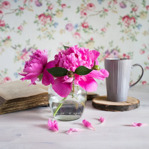 Fresh bouquet of peonies in a glass jar on a white wooden table. book and a cup of tea