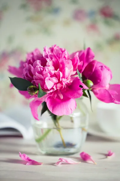 Fresh bouquet of peonies in a glass jar on a white wooden table. book and a cup of tea