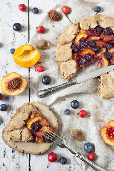 Wholemeal french galette with fruits sliced apricots peaches and blueberries on vintage rustic background with fresh fruits and rosehip on wooden table