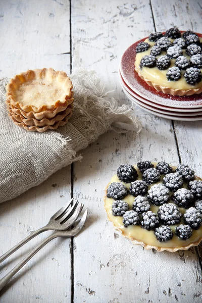 Two homemade blackberries tart with pastry cream on wooden table with cloth and fork