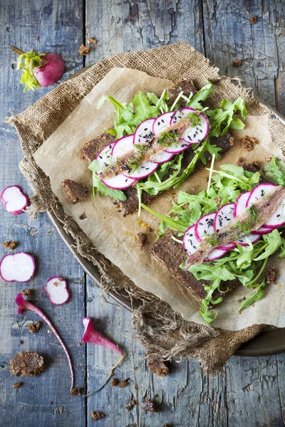 Two raw snack with slices of rye bread with rocket, radish slices, anchovies and seed on rustic table