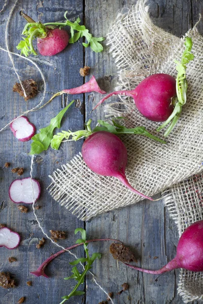 Raw whole and sliced radishes on burlap on rustic blue wooden table with leafs and crumbs