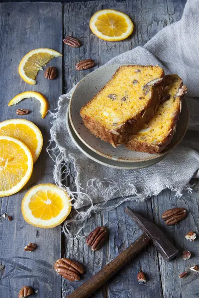 Two slices of citrus cake on plate on table with pecan walnuts and orange slices and little hammer