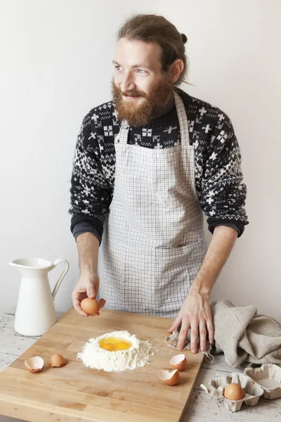 Young bearded stylish man in the kitchen with apron making dough for pasta with white flour and eggs