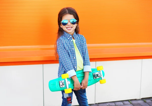 Happy stylish little girl child with skateboard over orange back