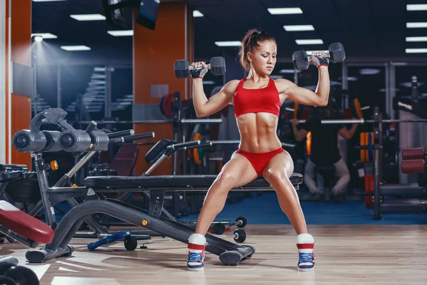 Sexy young athletics girl doing dumbbells press exercises. Fitness muscled woman in red sport clothing workout on bench in gym