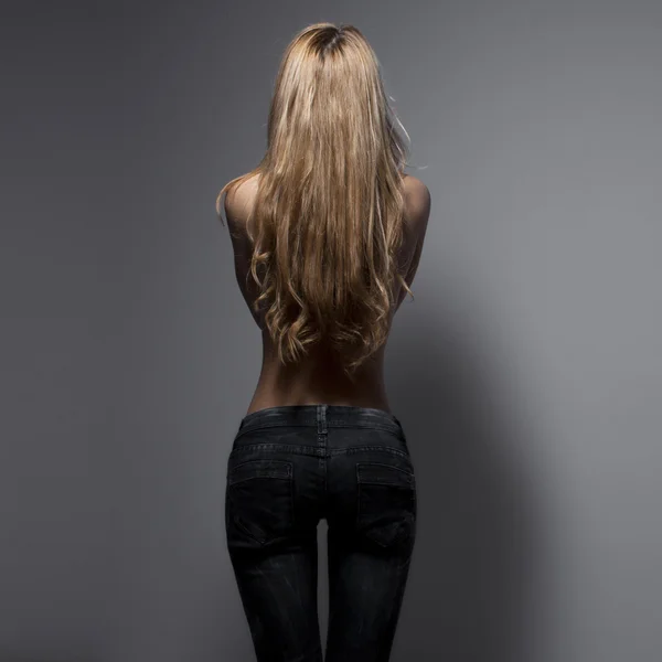 Fashion photo of a young sensual woman in jeans isolated on a grey background. Back posing. Blonde in jeans. Perfect body