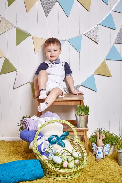 Beautiful little baby boy sitting on a chair with a knitted blanket Easter basket with colored eggs hay, Easter Bunny, a holy religious holiday, a happy guy cute child funny kid