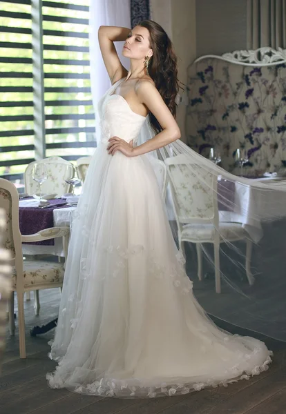 Beautiful young bride with long brown wavy hair in a lush white wedding dress of tulle, embroidered with beads corset, bow tied at the waist photographed in the interior of the restaurant