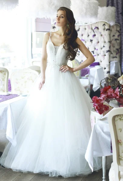Beautiful young bride with long brown wavy hair in a lush white wedding dress of tulle, embroidered with beads corset, bow tied at the waist photographed in the interior of the restaurant