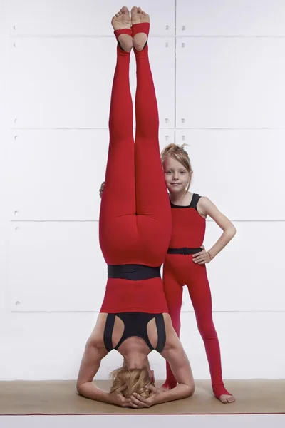 Mother and daughter doing yoga exercise, fitness, gym wearing the same comfortable tracksuits, family sports, sports paired woman standing up on head little girl holding her on legs they are on red
